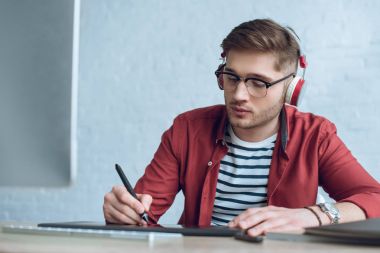 Freelancer man in headphones drawing with graphic tablet by table with computer clipart
