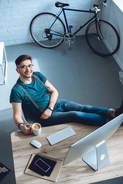 high angle view of handsome young freelancer smiling at camera while sitting at workplace 