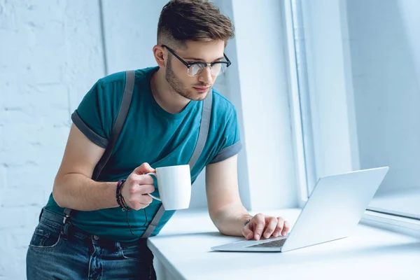 Νεαρός Άνδρας Γυαλιά Κρατώντας Φλιτζάνι Καφέ Και Χρησιμοποιούν Φορητό Υπολογιστή — Φωτογραφία Αρχείου