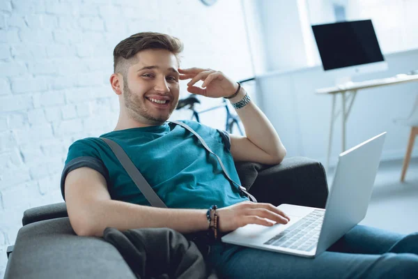 Joven Guapo Sonriendo Cámara Mientras Usa Ordenador Portátil Sofá Casa — Foto de Stock