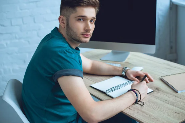 Guapo Joven Freelancer Tomando Notas Mirando Cámara Mientras Trabaja Casa —  Fotos de Stock