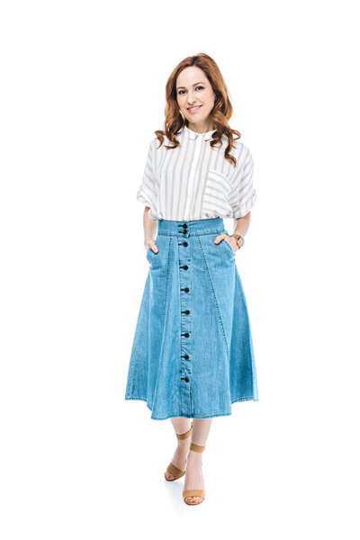 Вид в полный рост красивой женщины, стоящей с руками в карманах джинсовой юбки и улыбающейся камере, изолированной на белом фоне

