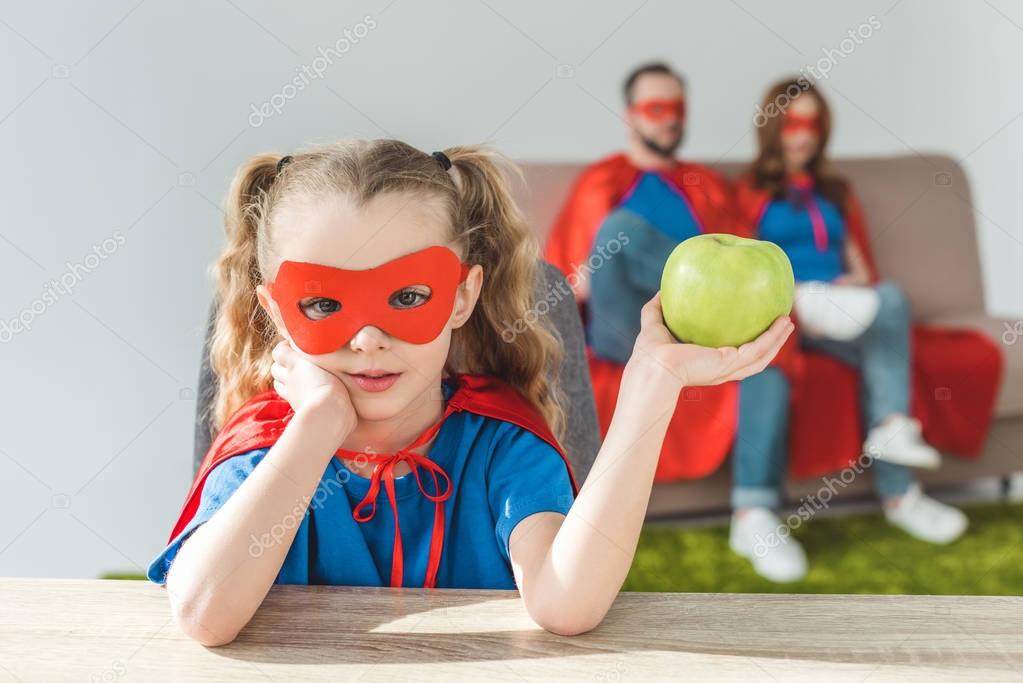 Girl in superhero costume holding apple and looking at camera while super parents sitting behind
