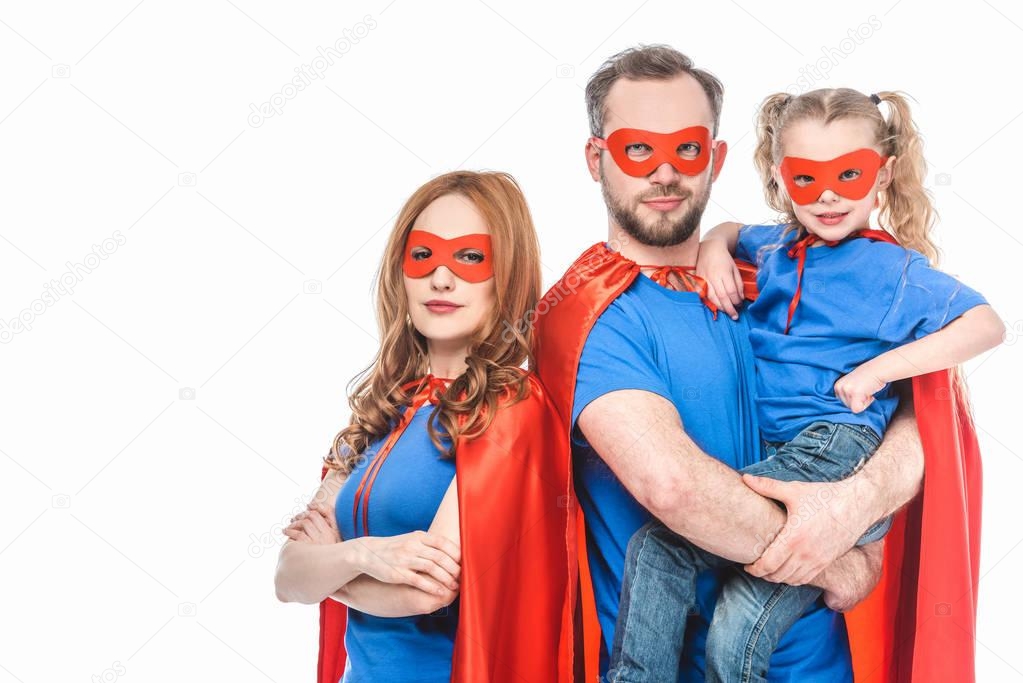 super parents with little daughter wearing masks and cloaks and looking at camera isolated on white