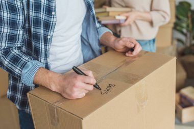 cropped shot of man signing cardboard box with wife with books in hands near by, moving home concept