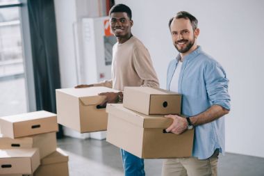 multiethnic coworkers holding cardboard boxes and smiling at camera during relocation   clipart