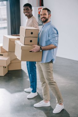 young multiethnic men holding cardboard boxes and smiling at camera during relocation  clipart