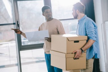 young multiethnic men looking at each other while holding paper and cardboard boxes during relocation clipart