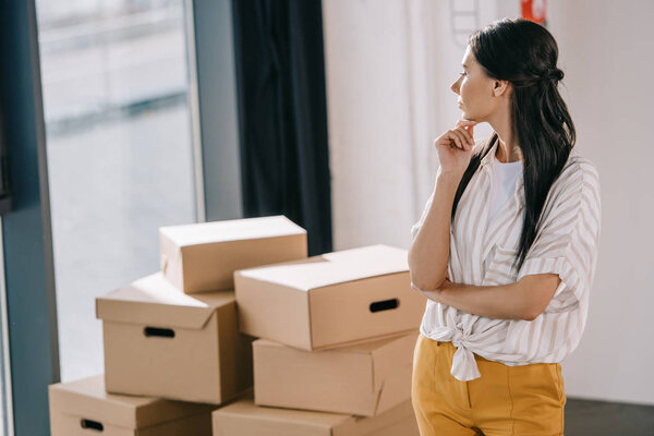 pensive young woman standing with hand on chin and looking at cardboard boxes in new office