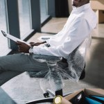Aus der Vogelperspektive: afrikanisch-amerikanischer Geschäftsmann hält Klemmbrett in der Hand und lächelt in die Kamera, während er während des Umzugs im neuen Büro sitzt