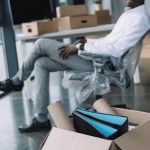 Close-up view of cardboard box with office supplies and african american businessman sitting behind in new office