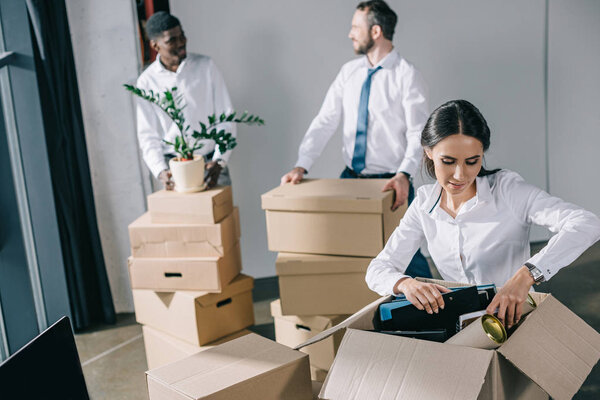 young businesswoman unpacking cardboard box and male colleagues standing behind in new office 