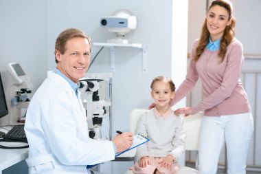 Anne, kızı ve kamera düşünülmeli danışmanlık odasında bakarak göz doktoruna gülümseyen