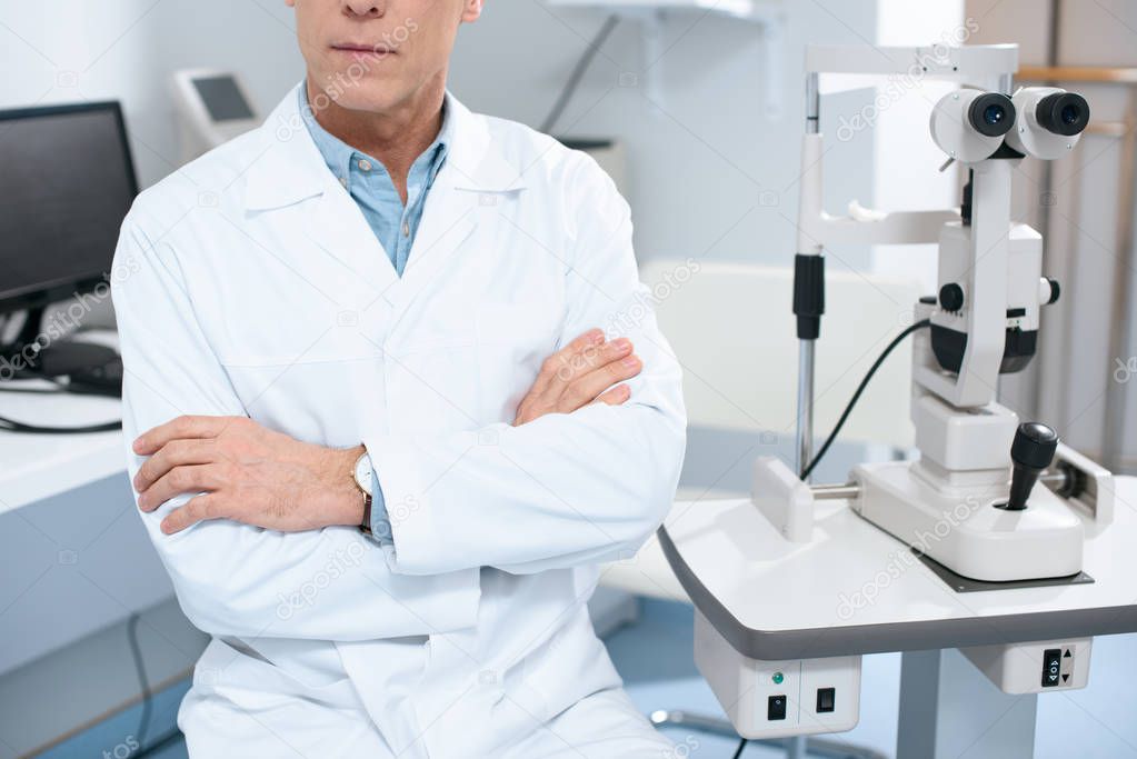 cropped image of ophthalmologist sitting with crossed arms near slit lamp in consulting room