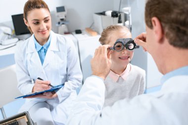 ophthalmologists examining kid eyes with trial frame and writing diagnosis clipart