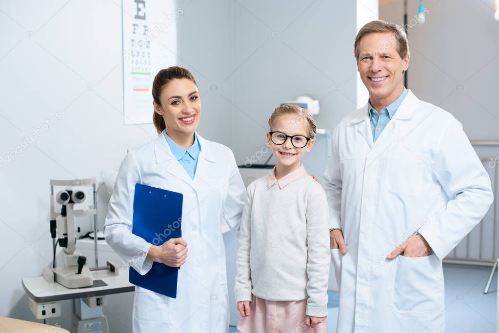 two smiling ophthalmologists and little child in glasses standing in clinic