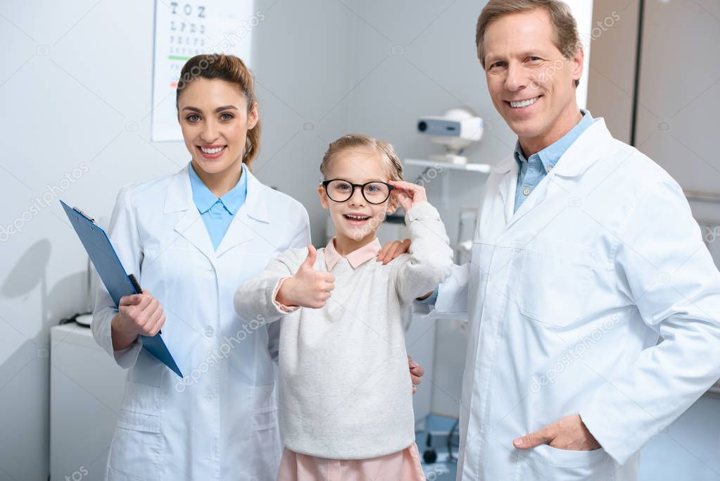 two smiling optometrists and little kid in eyeglasses showing thumb up