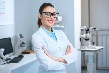 attractive professional ophthalmologist in glasses standing with crossed arms in optical clinic clipart