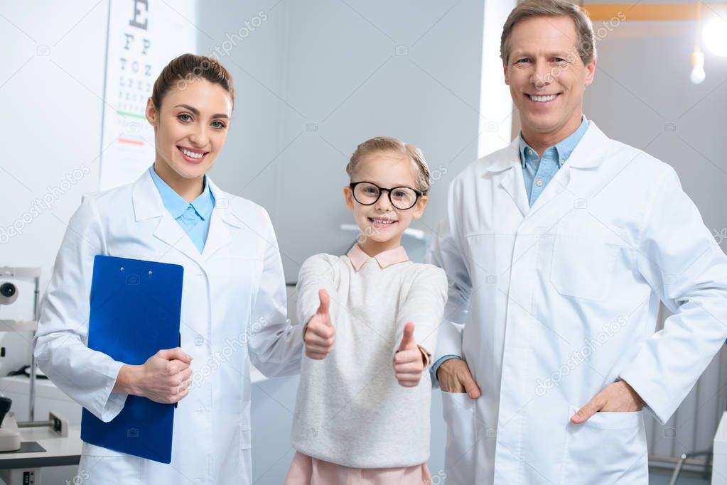 smiling optometrists and little kid in eyeglasses showing thumbs up
