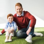 Happy redhead father and daughter sitting on grass and smiling at camera on grey