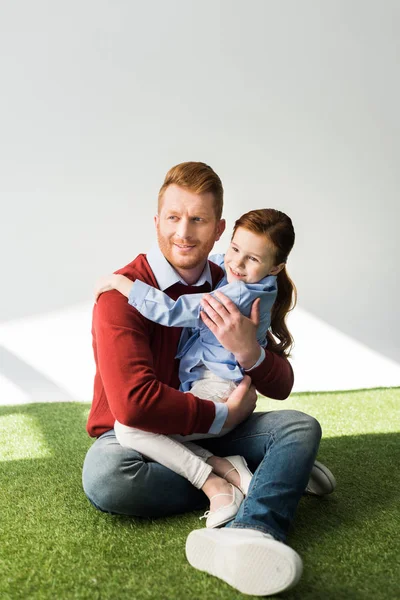 happy father and daughter hugging and looking away while sitting on grass