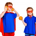Cute super children in masks and cloaks holding oranges isolated on white