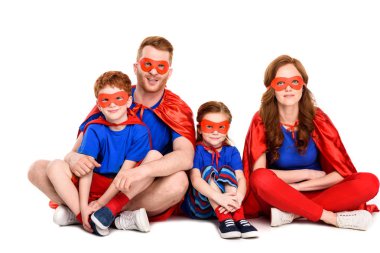 super family in costumes sitting together and smiling at camera isolated on white clipart
