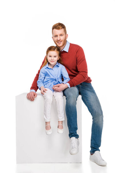 happy redhead father and daughter sitting on cube and smiling at camera isolated on white