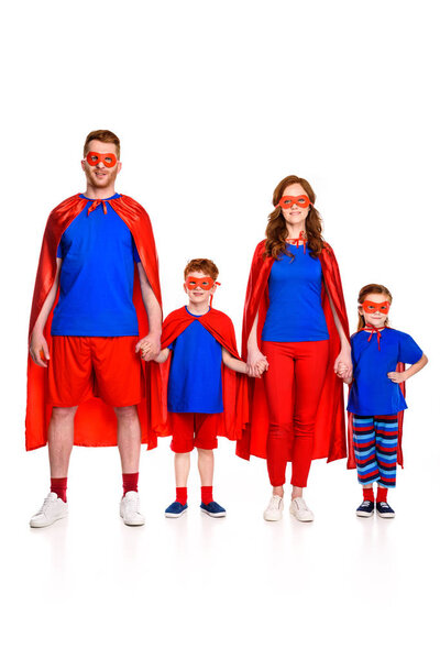 happy super family in masks and cloaks holding hands and smiling at camera isolated on white