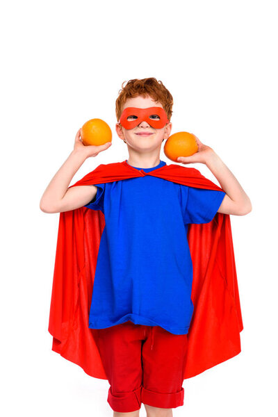 happy child in superhero costume holding oranges and smiling at camera isolated on white