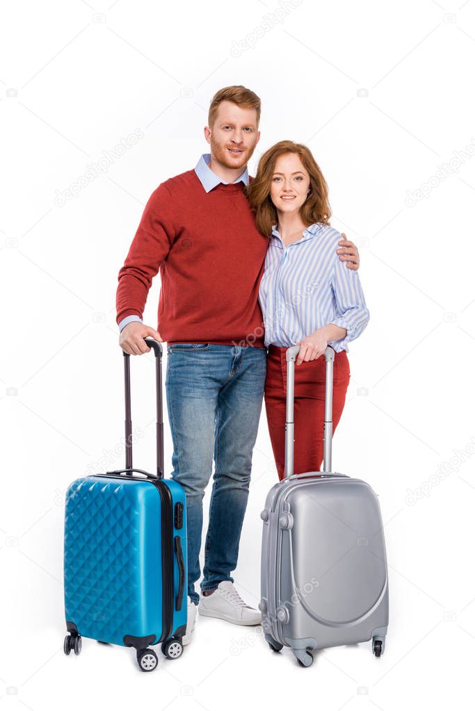 happy couple standing with suitcases and smiling at camera isolated on white