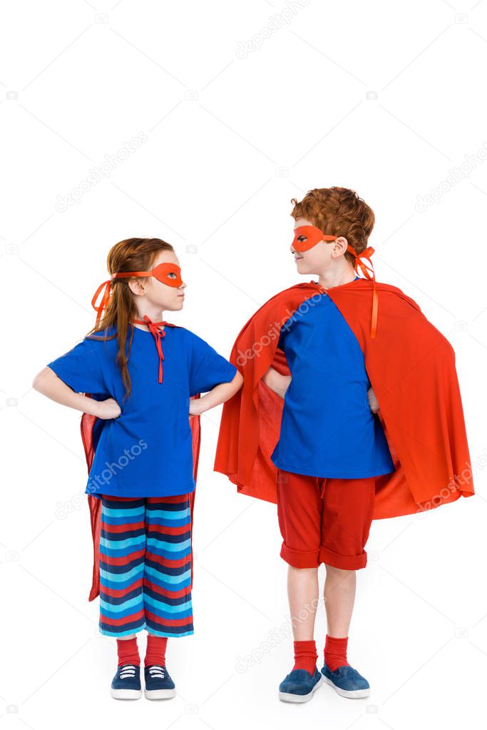 super kids in masks and cloaks standing with hands on waist and looking at each other isolated on white