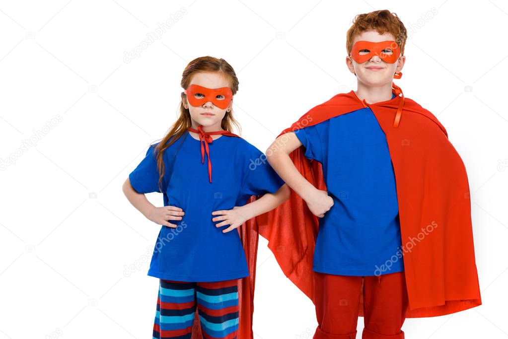 super kids in masks and cloaks standing with hands on waist and looking at camera isolated on white 