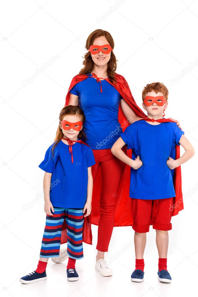 happy mother with cute little kids in superhero costumes standing together and smiling at camera isolated on white