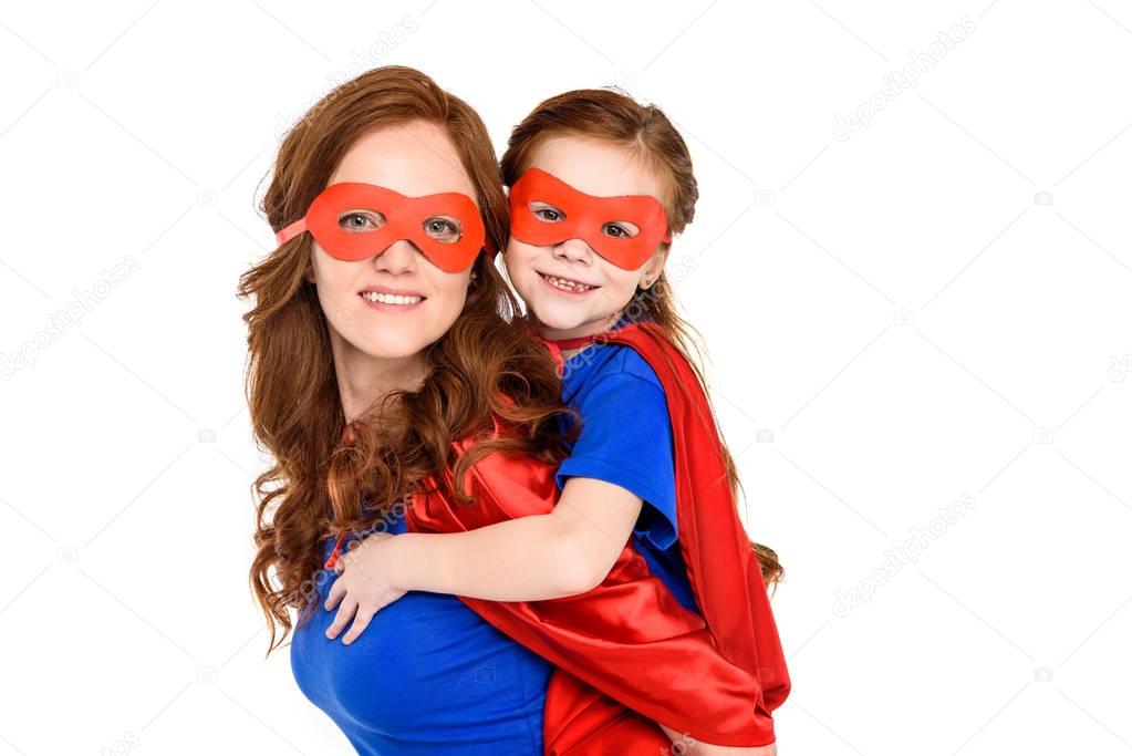 super mother piggybacking adorable daughter in mask and cloak and smiling at camera isolated on white