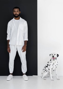 şık Afro-Amerikan yakışıklı beyaz giysiler ve Dalmaçya köpeği ayakta siyah beyaz duvarının yakınında