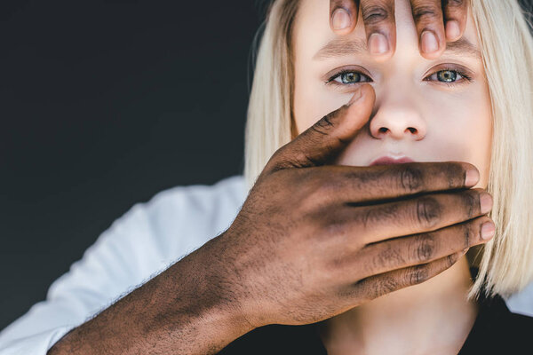 cropped image of african american boyfriend closing blonde girlfriend mouth isolated on black