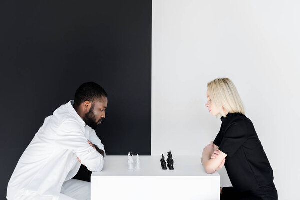side view of african american boyfriend and blonde girlfriend playing chess near black and white wall