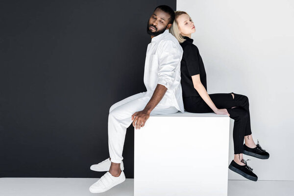 african american boyfriend and blonde girlfriend sitting back to back on white cube