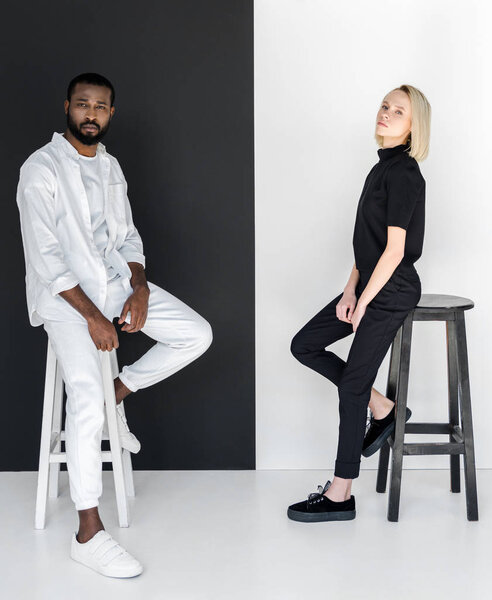 side view of african american boyfriend and blonde girlfriend sitting on chairs near black and white wall