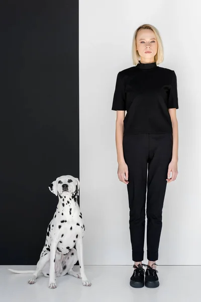 有吸引力的时尚金发女人穿着黑色的衣服站在黑色和白色的墙边与达尔马提亚狗 — 免费的图库照片