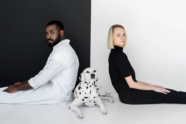 side view of multicultural couple sitting back to back with dalmatian dog on floor