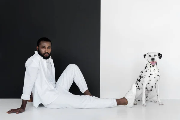 英俊时尚的非洲裔美国人穿着白色衣服和达尔马提亚狗坐在黑色和白色的墙壁附近 — 图库照片