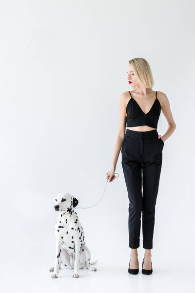 attractive stylish blonde woman in black clothes holding dalmatian dog leash on white