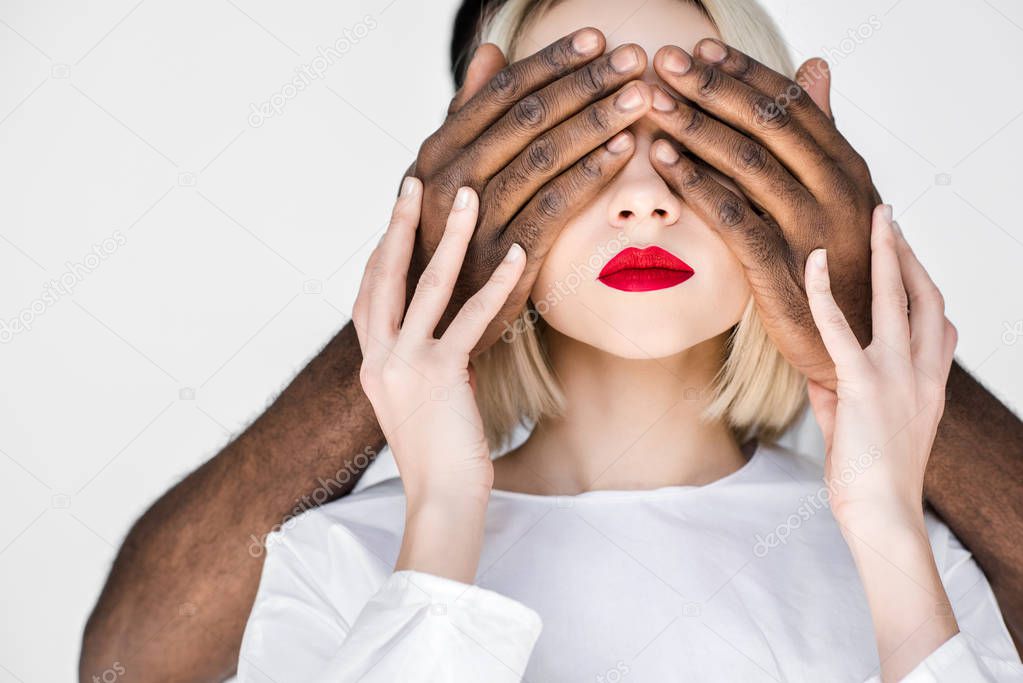 cropped image of african american boyfriend closing eyes of blonde girlfriend isolated on white