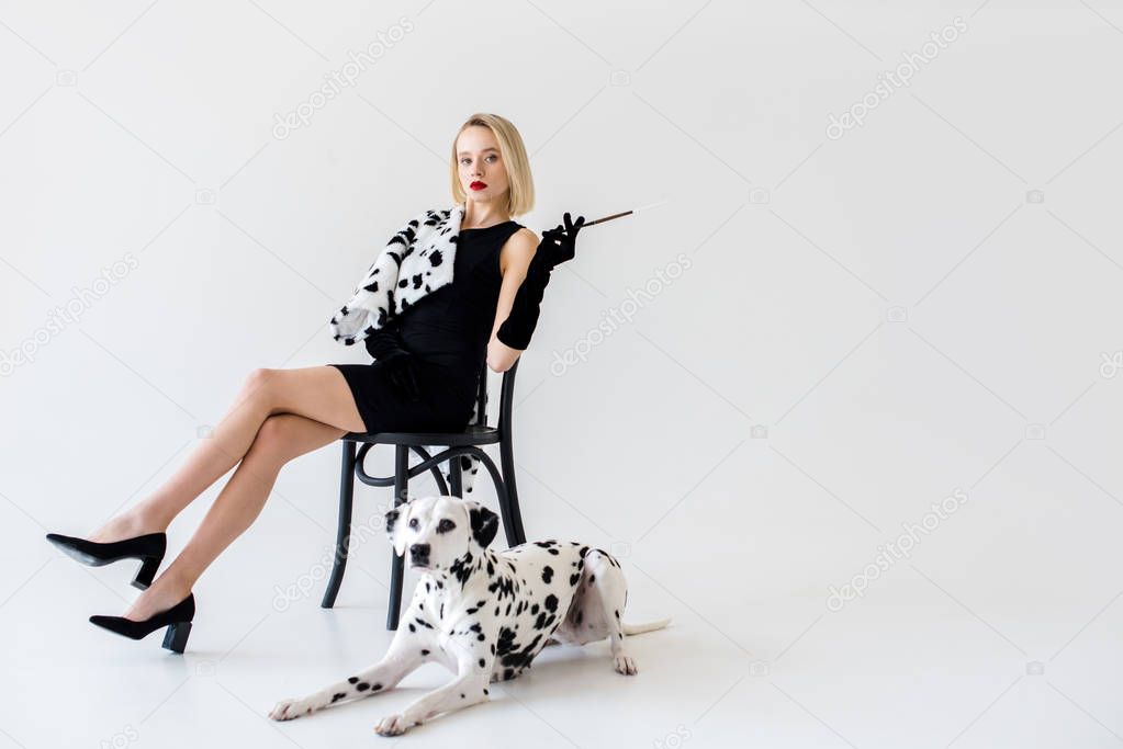 attractive stylish blonde woman in black dress sitting on chair, dalmatian dog lying on floor