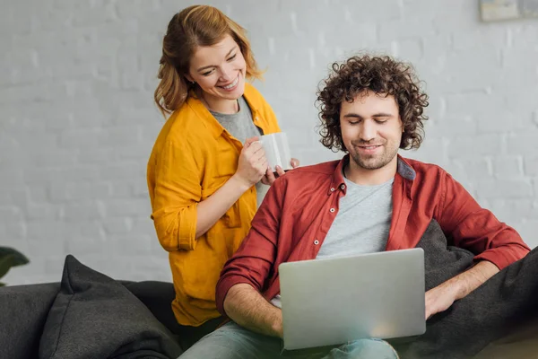 Sonriente Joven Mujer Sosteniendo Taza Mirando Novio Usando Ordenador Portátil — Foto de Stock