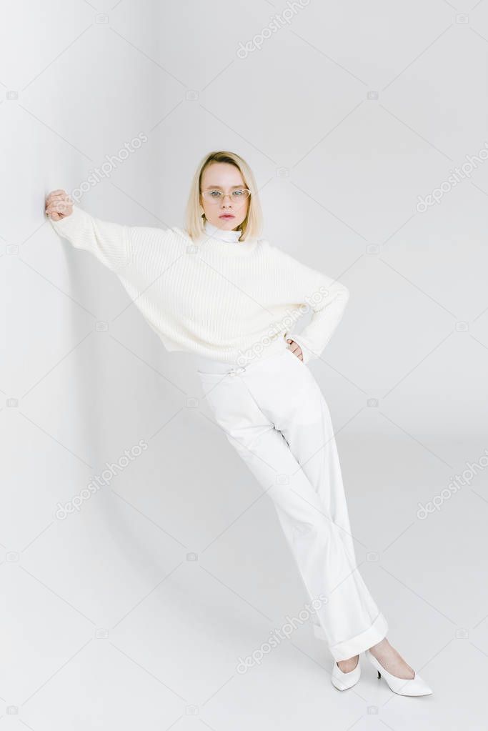 beautiful stylish blonde woman in white clothes leaning on white wall and looking at camera