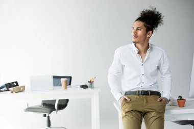 handsome young african american businessman with hands in pockets sitting on table and looking away in office clipart