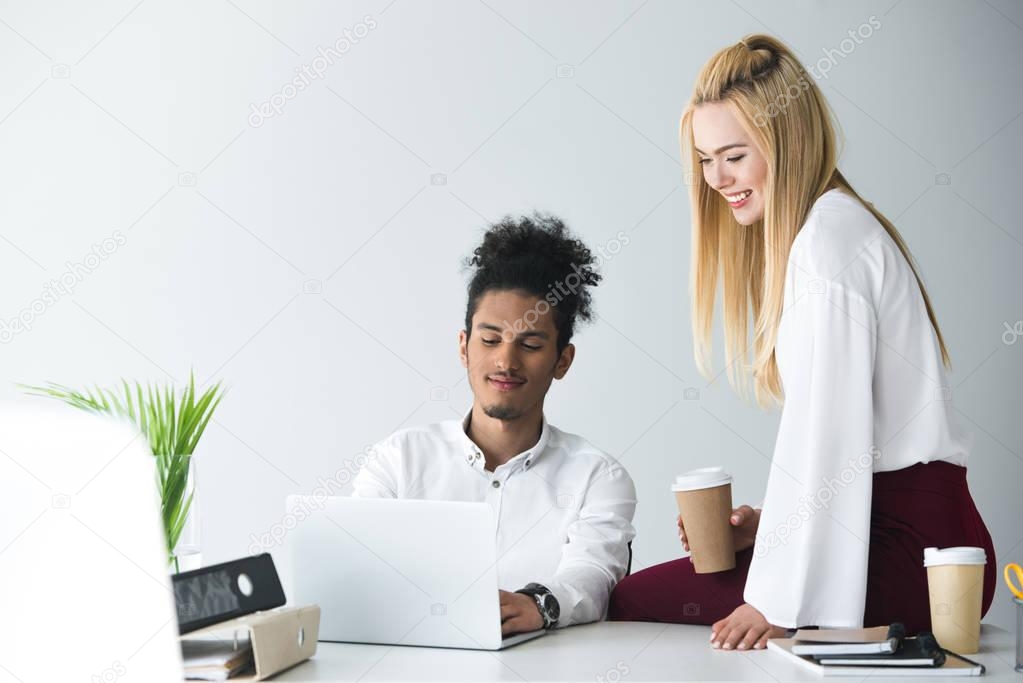 smiling young businesswoman holding paper cup and looking at businessman using laptop in office 
